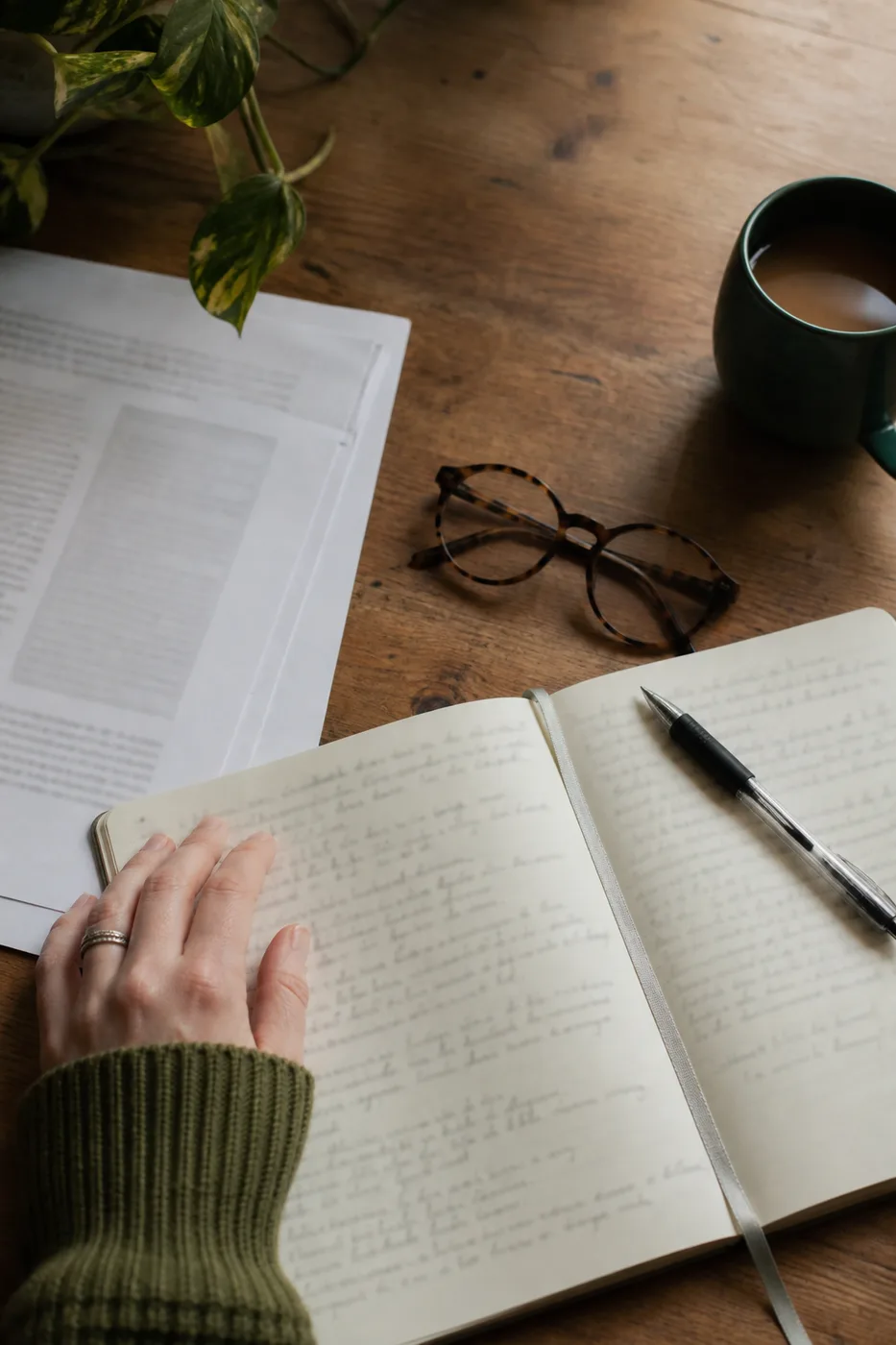 An open notebook with handwriting, printed research papers, reading glasses and a mug on a desk